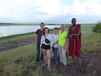 Eine Gruppe von fünf Personen steht auf einem Hügel mit Blick auf eine grüne Landschaft und einen ruhigen See im Hintergrund.