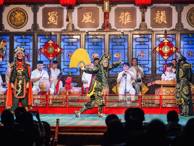 Auf einer Bühne führen zwei Schauspieler in traditioneller Kleidung eine lebhafte Sichuan-Oper auf, umgeben von Musikern.