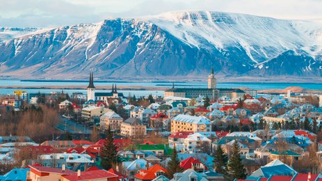 Blick auf Reykjavik mit bunten Häusern, umgeben von schneebedeckten Bergen und einem ruhigen Wasser im Hintergrund.