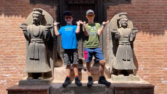 Zwei Jungen posieren fröhlich zwischen zwei beeindruckenden Steinfiguren in Bhaktapur, umgeben von rotem Ziegelmauerwerk.