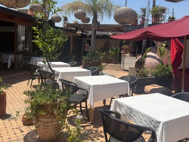 Eine sonnige Terrasse mit weißen Tischen und Stühlen, umgeben von grünen Pflanzen und geflochtenen Lampen, lädt zum Verweilen ein.