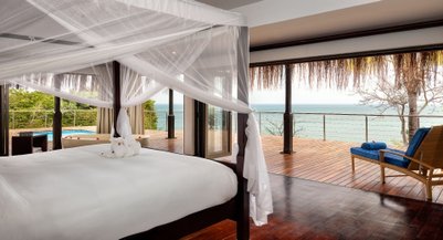 Ein luxuriöses Schlafzimmer mit einem großen Bett, umgeben von einem Moskitonetz und Blick auf das Meer durch große Fenster.