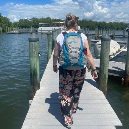 Eine Person mit einem Rucksack geht auf einem Steg entlang, umgeben von Wasser und Booten unter einem strahlend blauen Himmel.