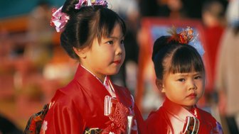 Zwei kleine Mädchen in traditioneller japanischer Kleidung, die mit Blumen im Haar an einem festlichen Ort stehen.
