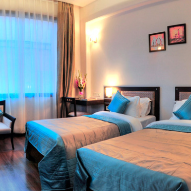 Zwei Einzelbetten mit blauen Bettüberwürfen stehen in einem hellen, modernen Hotelzimmer mit großen Fenstern.