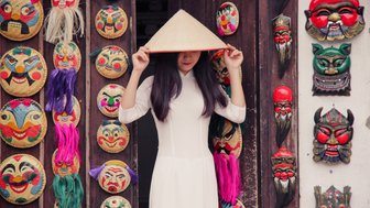 Eine Frau in traditioneller Kleidung steht vor einer Wand mit bunten Masken und trägt einen vietnamesischen Reisstrohhut.