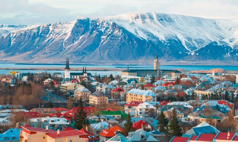 Blick auf Reykjavik mit bunten Häusern, umgeben von schneebedeckten Bergen und einem ruhigen Wasser im Hintergrund.