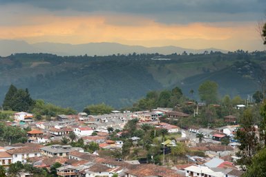 Eine malerische Aussicht auf ein Dorf, umgeben von sanften Hügeln und bewaldeten Bergen unter einem bewölkten Himmel.