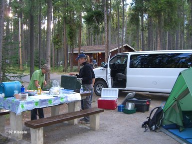 Zwei Personen bereiten ein Picknick an einem Tisch im Wald vor, umgeben von hohen Bäumen und einem Wohnmobil.