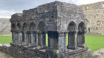 Ein historisches Steingebäude mit Bögen und Säulen, umgeben von grünem Gras und alten Mauern im Hintergrund.