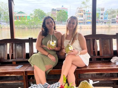 Zwei Frauen sitzen auf einem Boot im Mekong-Delta und genießen frische Früchte und Getränke, umgeben von einer malerischen Kulisse.
