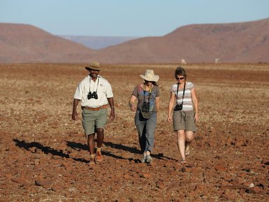 Drei Wanderer erkunden die trockene Landschaft von Damaraland, umgeben von roten Felsen und sanften Hügeln im Hintergrund.