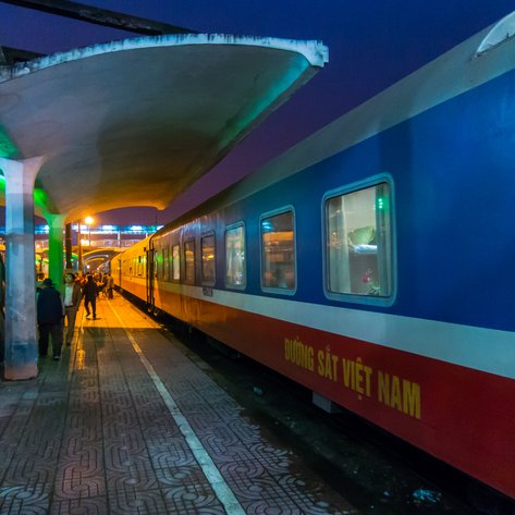Ein Nachtzug steht an einem beleuchteten Bahnhof, während Passagiere auf dem Bahnsteig warten und sich unterhalten.