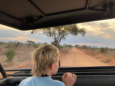 Ein Junge schaut aus einem Geländewagen auf eine staubige Straße, umgeben von Bäumen und einer weiten Landschaft.