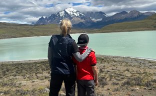Eine Frau und ein Junge stehen am Ufer eines türkisfarbenen Sees und blicken auf die majestätischen Berge im Hintergrund.