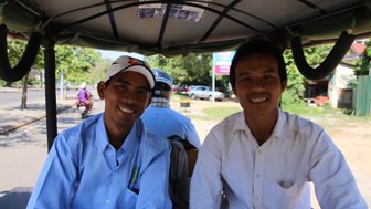 Zwei Männer lächeln fröhlich in einem Tuk-Tuk, während sie durch eine belebte Straße fahren, umgeben von Bäumen und Fahrzeugen.