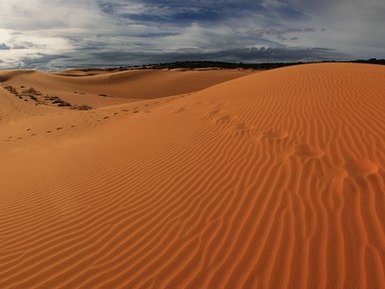 Die sanften, orangefarbenen Sanddünen erstrecken sich unter einem dramatischen Himmel mit Wolken und Lichtspielen.