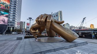 Eine große, goldene Statue einer Hand, die ein Schild mit der Aufschrift 'Gangnam Style' hält, steht in einer urbanen Umgebung.
