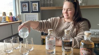 Eine Frau mit langen Haaren gießt klaren Alkohol in ein Glas, während sie an einer Holztheke steht.
