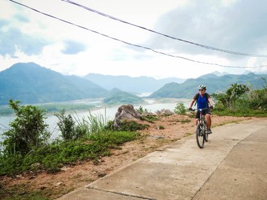Ein Radfahrer fährt auf einem schmalen Weg mit Blick auf die Berge und den Fluss im Hintergrund, umgeben von üppigem Grün.