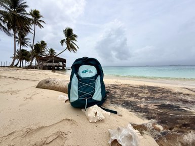 Ein türkisfarbener Rucksack steht im Sand am Strand, mit einer gemütlichen Holzhütte und Palmen im Hintergrund.