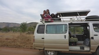 Eine Gruppe von Kindern und Erwachsenen sitzt fröhlich auf dem Dach eines Vans, umgeben von einer ländlichen Landschaft.