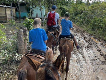 Drei Reiter, darunter zwei Kinder, reiten auf einem schlammigen Weg durch eine grüne Landschaft mit Bäumen.