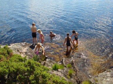 Eine Gruppe von Kindern spielt am Ufer eines klaren Sees, während einige im Wasser planschen und andere auf den Steinen stehen.
