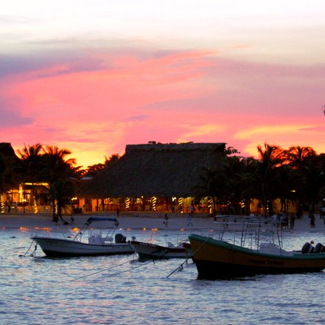 Ein malerischer Sonnenuntergang über einem ruhigen Gewässer, umgeben von Palmen und beleuchteten Restaurants am Strand.