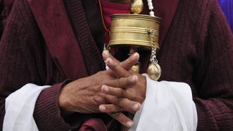 Eine ältere Person hält eine Gebetsmühle in den Händen, umgeben von traditioneller tibetischer Kleidung und einem weißen Tuch.