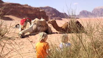 Zwei Kinder sitzen im Wadi Rum, während Kamele im Hintergrund auf dem Sand ruhen, umgeben von sanften Hügeln.