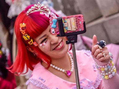 Eine fröhliche Frau mit rotem Haar und bunten Accessoires macht ein Selfie mit einem Smartphone in der Hand.