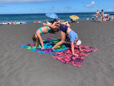 Zwei Mädchen machen akrobatische Übungen auf bunten Handtüchern am Strand, umgeben von anderen Sonnenanbetern.