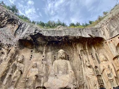 Imposante Buddha-Statue in einer Felsenkirche, umgeben von kunstvollen Reliefs und einer dramatischen Himmelskulisse.