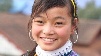 Ein fröhliches vietnamesisches Mädchen mit langen Haaren und großen Ohrringen lächelt in die Kamera, umgeben von einer ländlichen Kulisse.