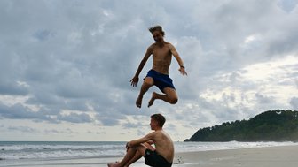 Ein Junge springt fröhlich in die Luft am Strand, während ein anderer entspannt am Ufer sitzt und das Meer beobachtet.