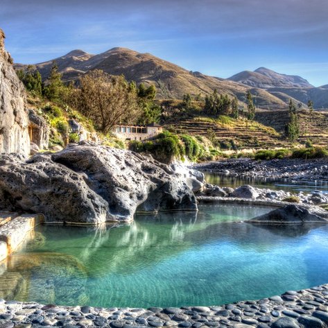 Ein klarer Thermalpool umgeben von Felsen und üppiger Vegetation, mit sanften Hügeln im Hintergrund.