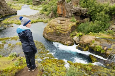 Eine Person steht am Rand einer Klippe und blickt auf einen klaren Fluss, umgeben von üppiger, grüner Vegetation.