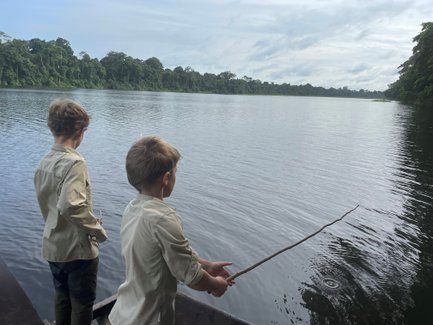 Zwei Jungen stehen am Ufer eines ruhigen Gewässers und versuchen, mit einem Stock zu fischen, umgeben von üppigem Grün.