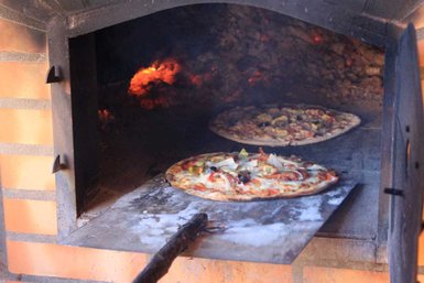 Zwei frisch gebackene Pizzen liegen auf einem Holzbrett in einem traditionellen Holzofen, umgeben von glühenden Kohlen.
