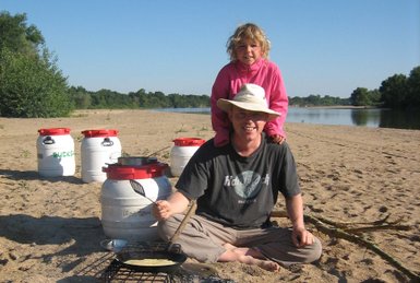 Ein Mann sitzt am Strand und kocht mit einer Pfanne über einem offenen Feuer, während ein Kind auf seinen Schultern sitzt.