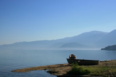 Ein ruhiger See mit sanften Wellen, umgeben von Bergen und einem kleinen Boot am Ufer, das in der Morgensonne glänzt.