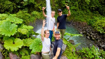 Eine Gruppe von vier Freunden posiert glücklich vor einem beeindruckenden Wasserfall in üppigem Grün.