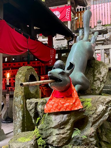 Eine bronzeartige Statue eines Hundes mit einem Stock im Mund, gekleidet in ein leuchtend rotes Tuch, umgeben von Steinen.