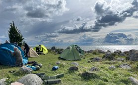 Eine Gruppe von Menschen baut Zelte am Ufer eines Sees auf, umgeben von großen Steinen und einer dramatischen Wolkenlandschaft.