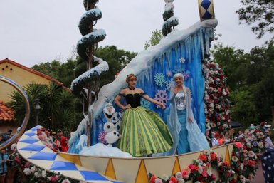 Anna und Elsa stehen auf einem festlich dekorierten Wagen, umgeben von bunten Blumen und schneebedeckten Elementen.