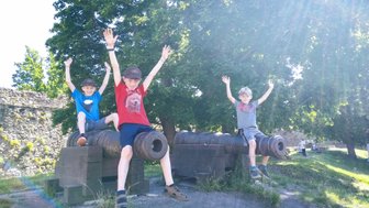 Drei fröhliche Kinder sitzen auf Kanonen und heben die Hände in die Luft, um ihre Freude auszudrücken.