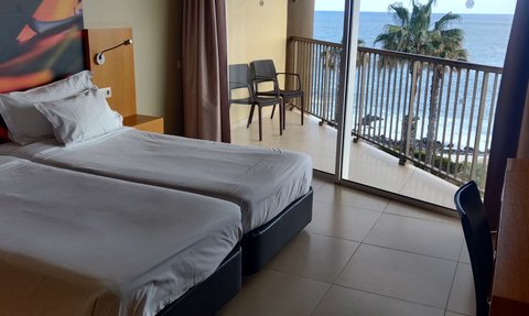 Ein helles Hotelzimmer mit zwei Einzelbetten, Blick auf das Meer und Palmen durch große Fenster.