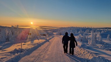 Ein Paar spaziert auf einem verschneiten Weg, während die Sonne hinter den Bergen untergeht und den Himmel in warmen Farben erleuchtet.