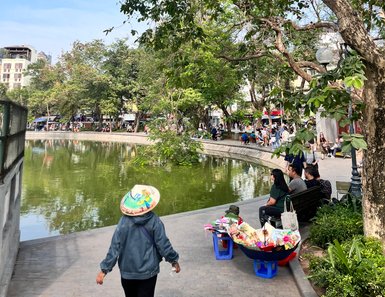 Eine Person mit einem traditionellen Hut geht entlang eines malerischen Sees, umgeben von üppigem Grün und fröhlichen Menschen.